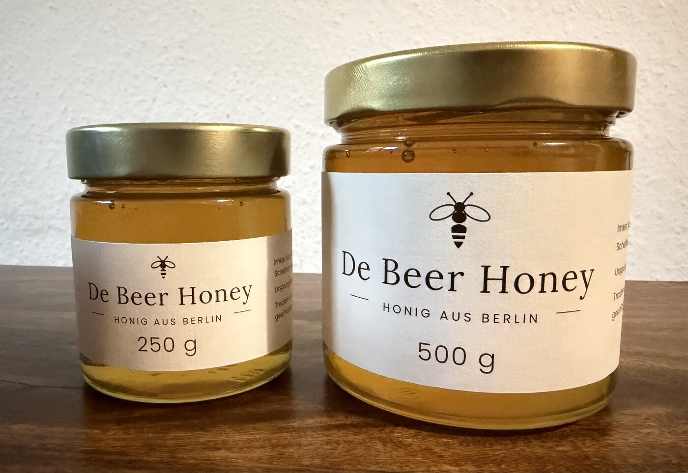 Zwei Gläser De Beer Honey mit 250 g und 500 g Inhalt auf einer Holzplatte